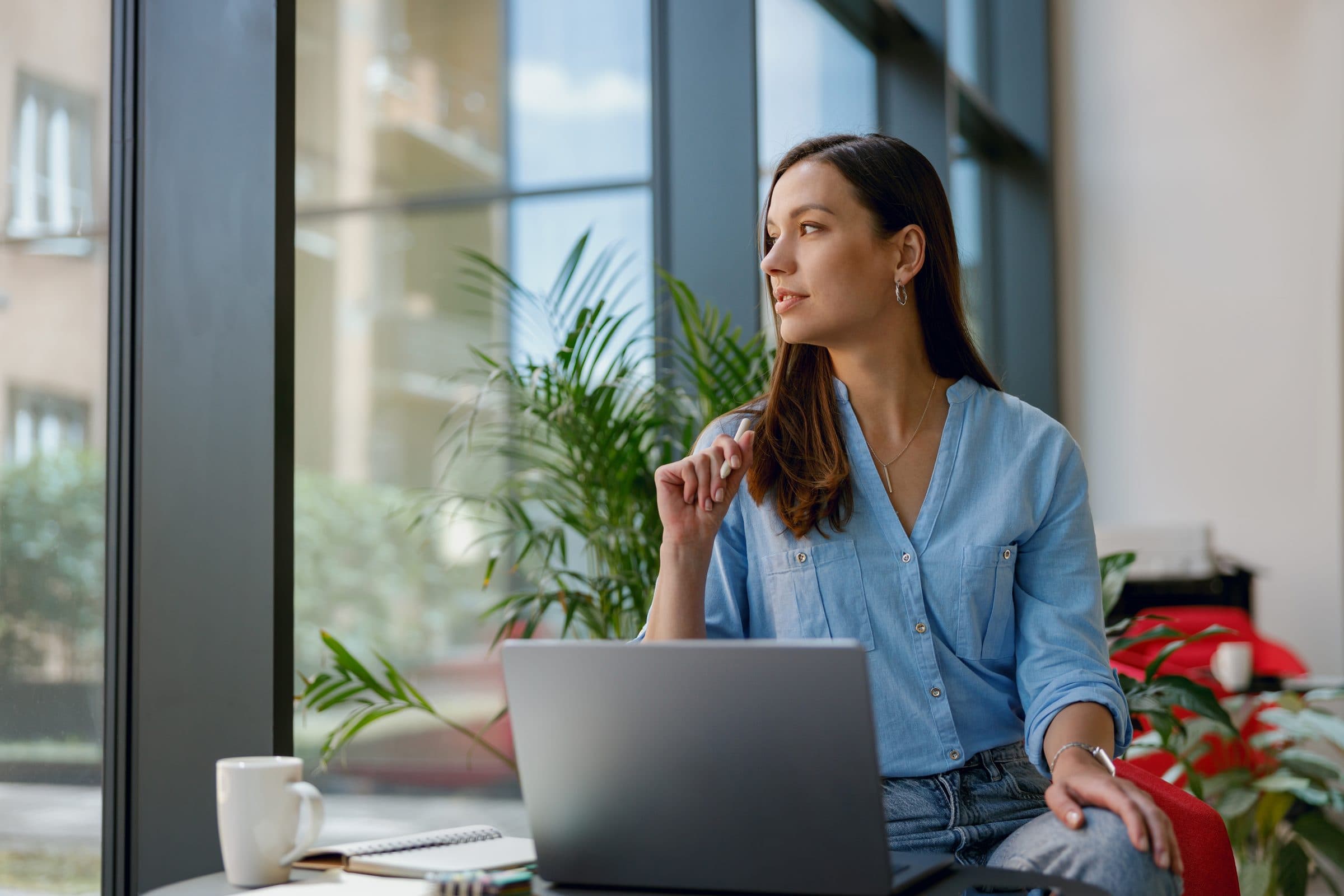 A woman in a blue shirt at her laptop by a tall window, looking outside