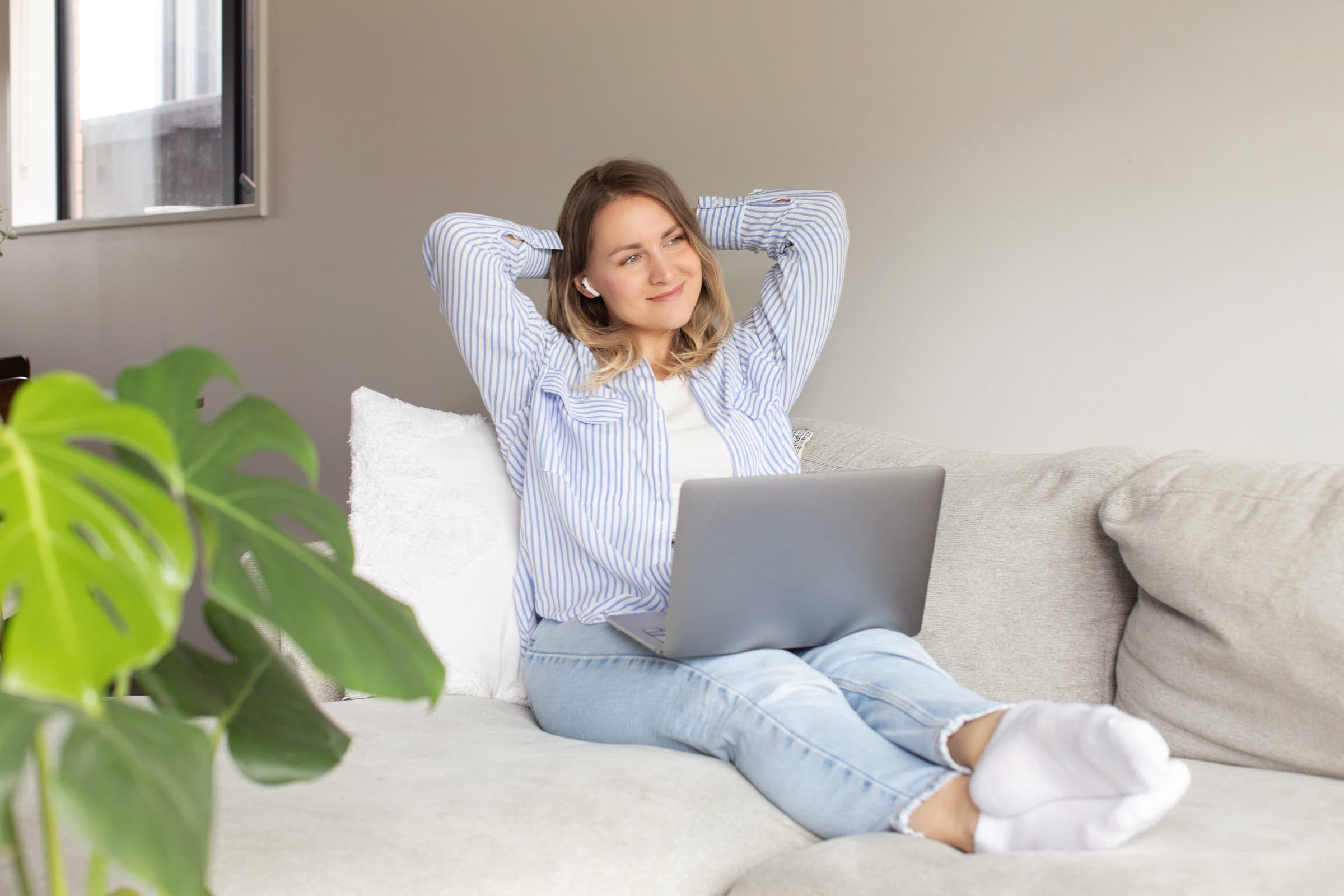 A woman relaxing on a sofa with a laptop on her lap, hands behind her head