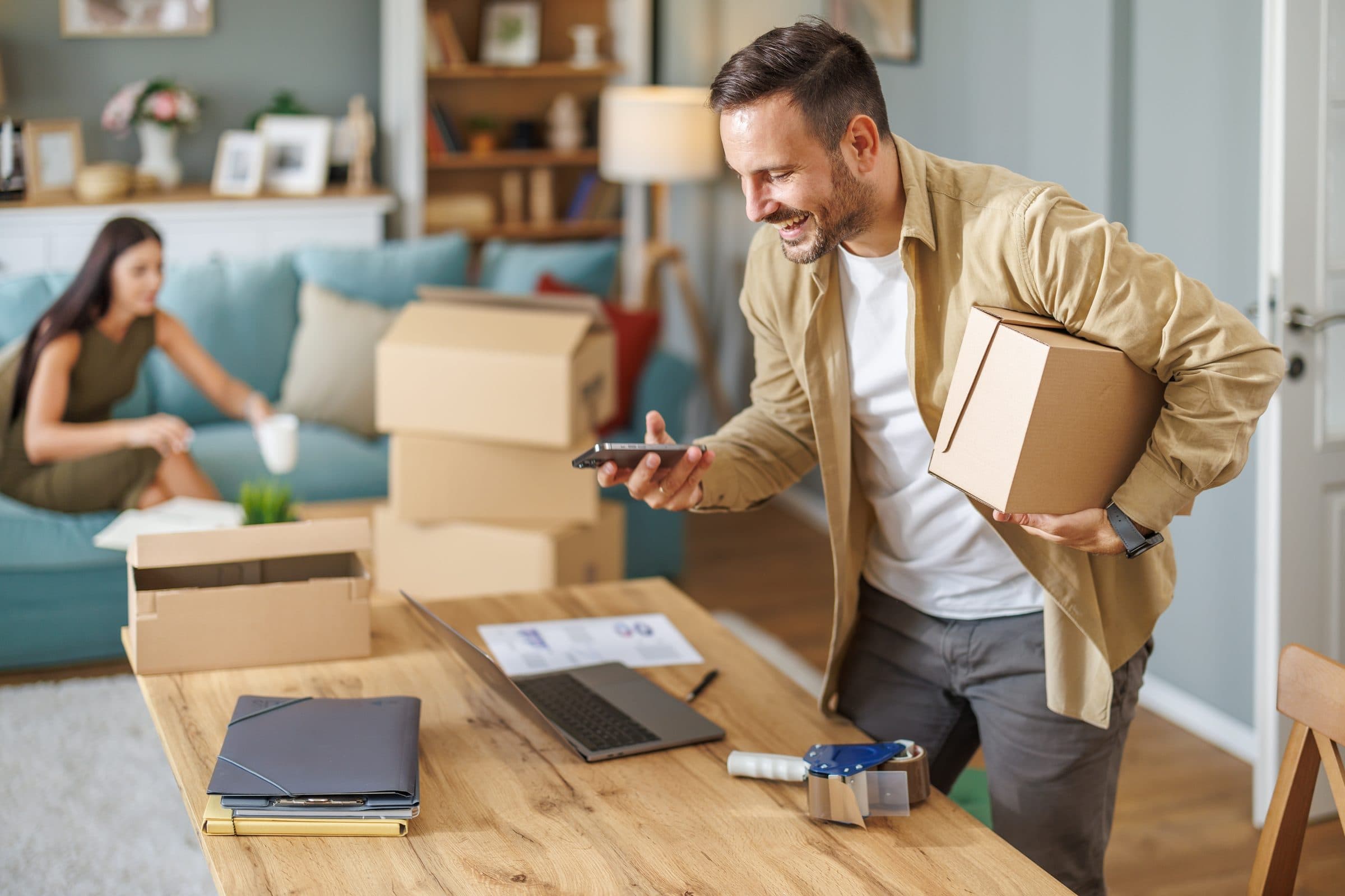 A man shipping an order: a parcel under one arm, photographing it with his phone beside a laptop